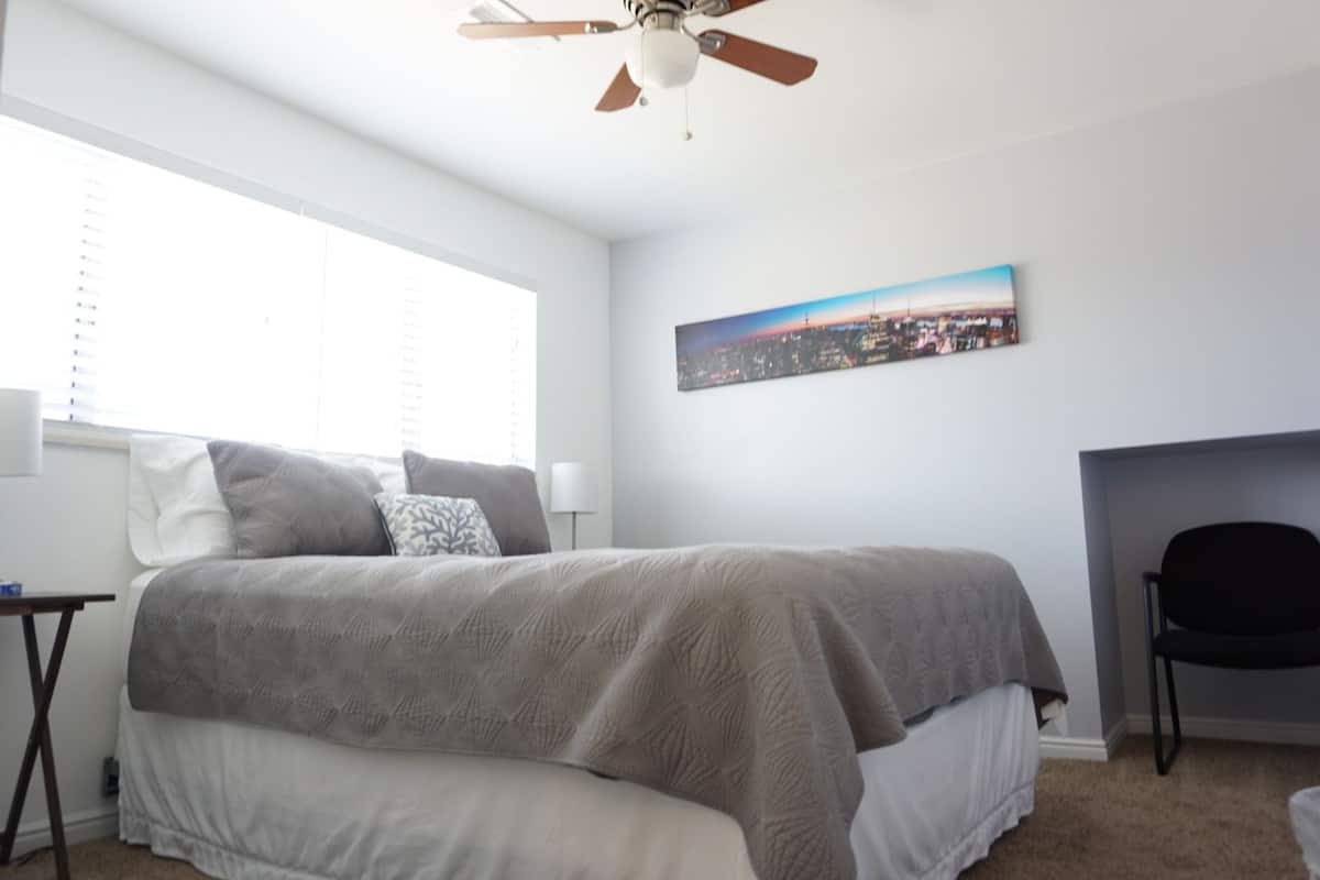 A bedroom with a large bed, a ceiling fan, a cityscape painting on the wall, and a black chair.