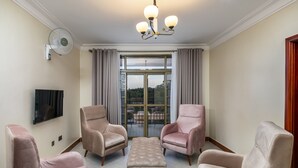Un salon comprenant deux fauteuils confortables, un pouf assorti, un téléviseur à écran plat, un ventilateur de plafond et une grande fenêtre avec des rideaux.