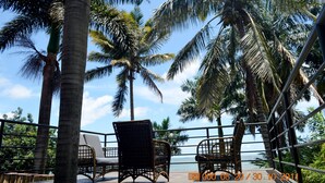 Un balcon avec des fauteuils en osier et une balustrade donnant sur une plage bordée de palmiers et l’océan.