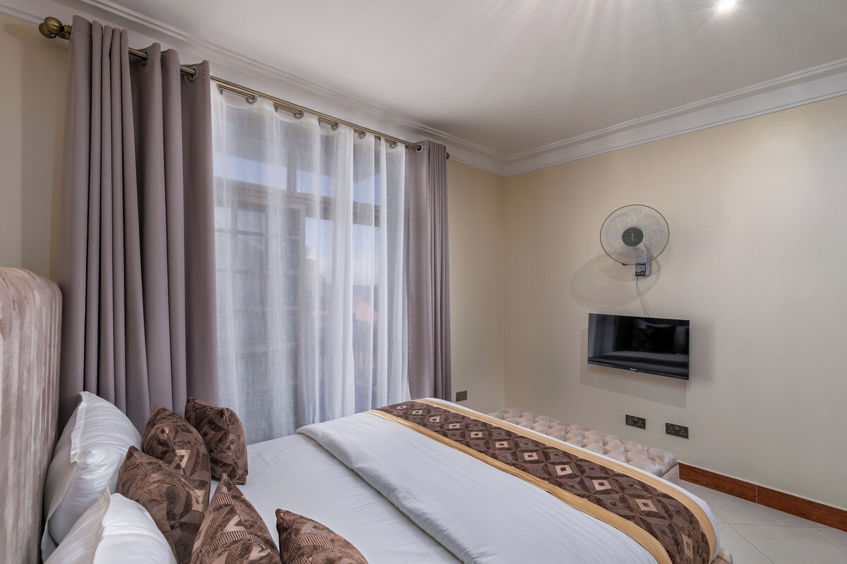 A hotel room with a large bed, a flat-screen TV mounted on the wall, a ceiling fan, and a window with curtains.