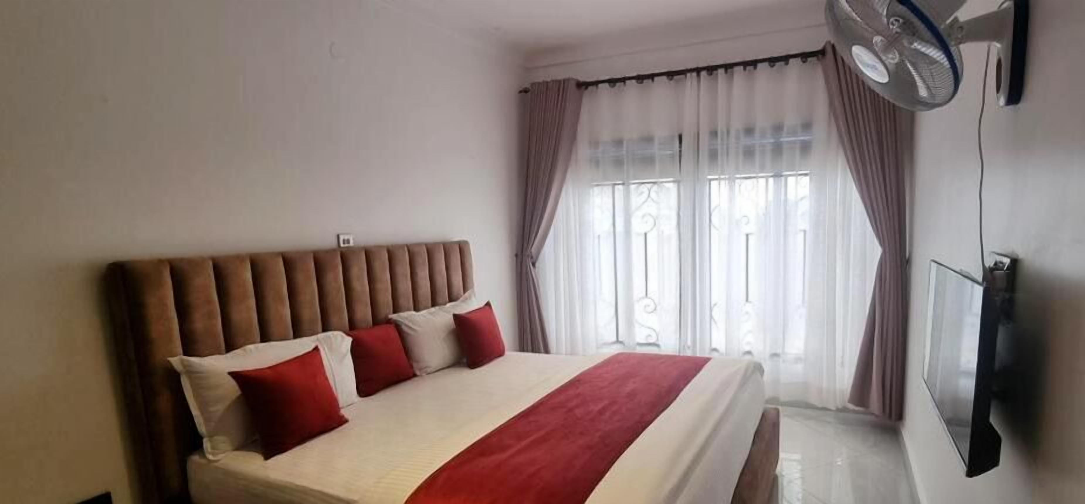 Ein Hotelzimmer mit einem großen Bett, einem flachbildfernseher an der Wand und einem Fenster mit durchsichtigen Vorhängen.