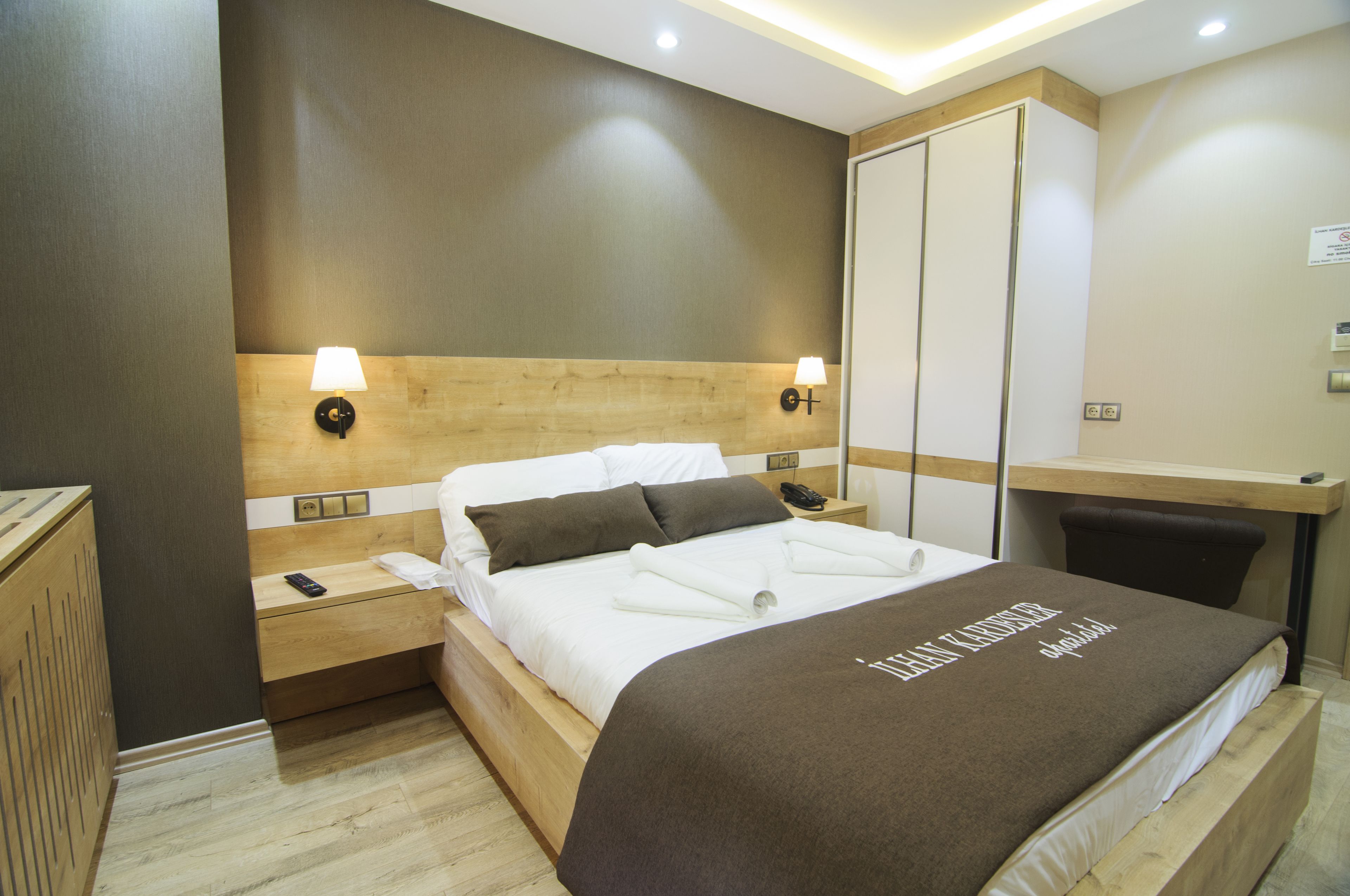 A modern hotel room with a wooden headboard, two bedside lamps, a neatly made bed with a brown blanket, and a wooden nightstand with a remote control.