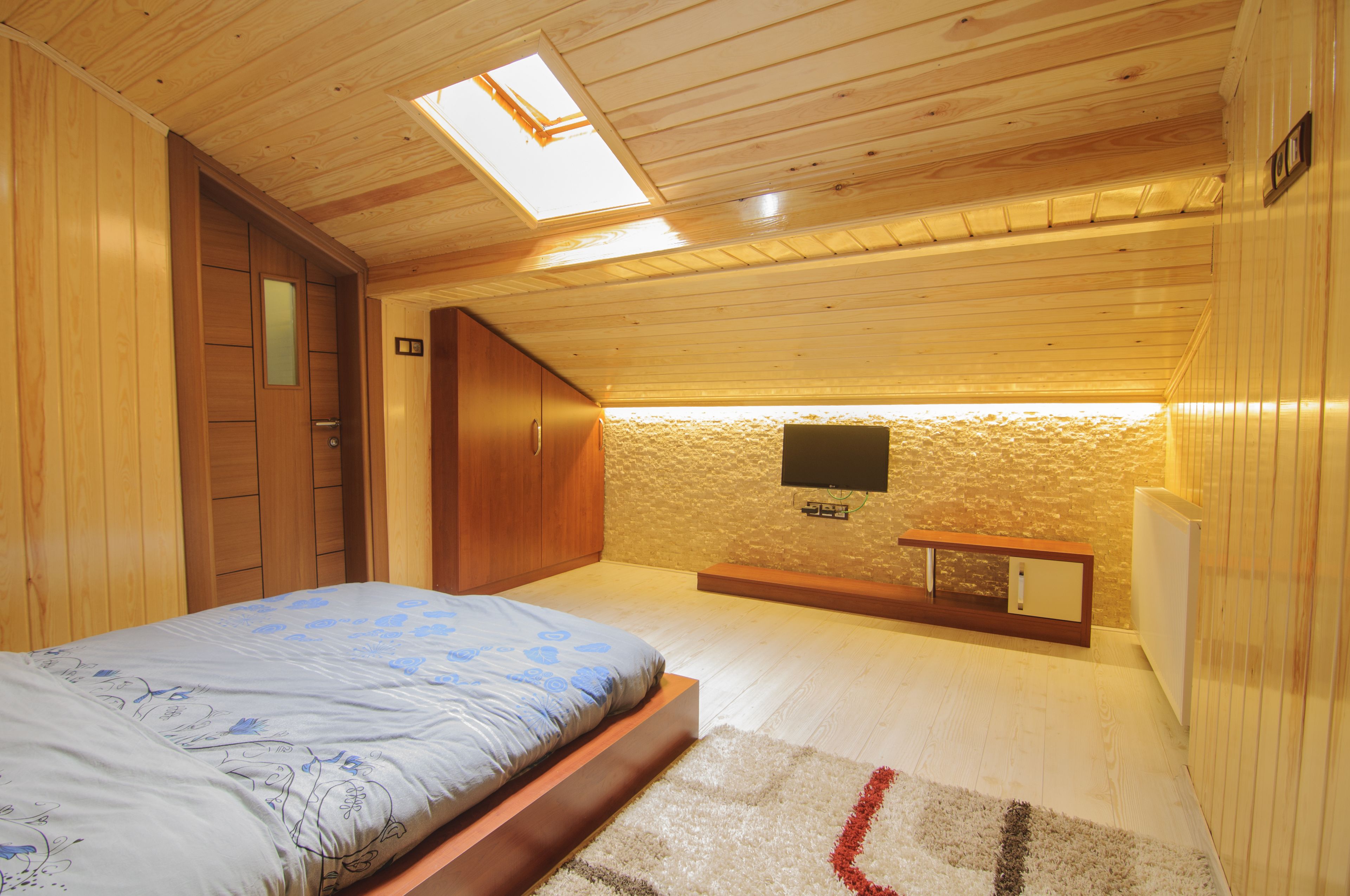 A bedroom with a bed, wooden walls, a skylight, a television, and a small bench.