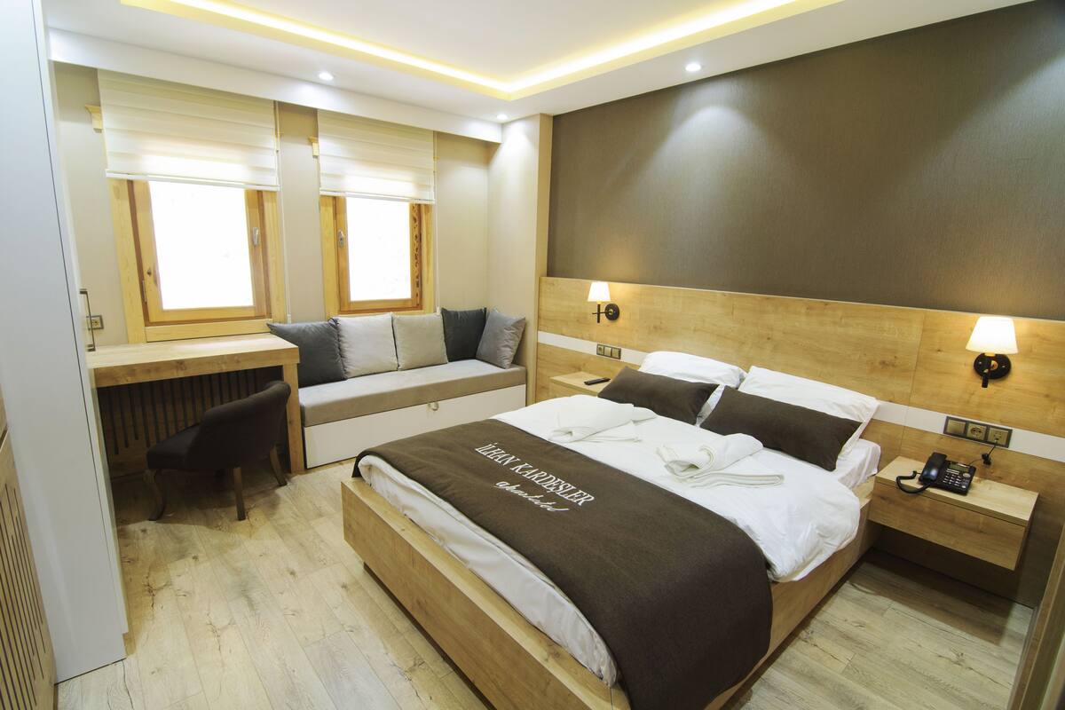 Ein Hotelzimmer mit einem großen Bett, einem Schreibtisch mit Stuhl, einem Nachttisch mit Lampe und einem Fenster mit Jalousien.