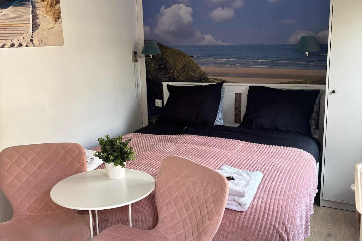 A bedroom with a bed, two pink chairs, a small table, and a potted plant.