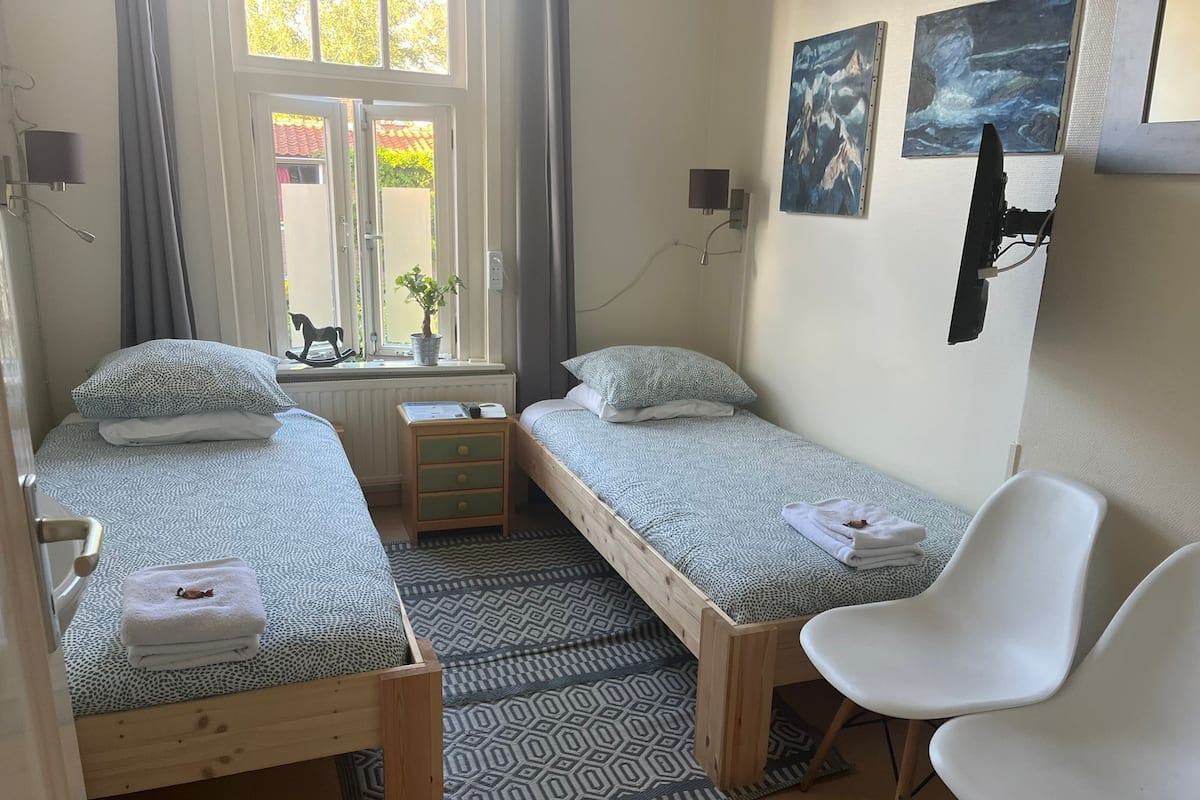 A small, well-lit bedroom with two single beds, a wooden nightstand, a white chair, and a window with a view of greenery outside.