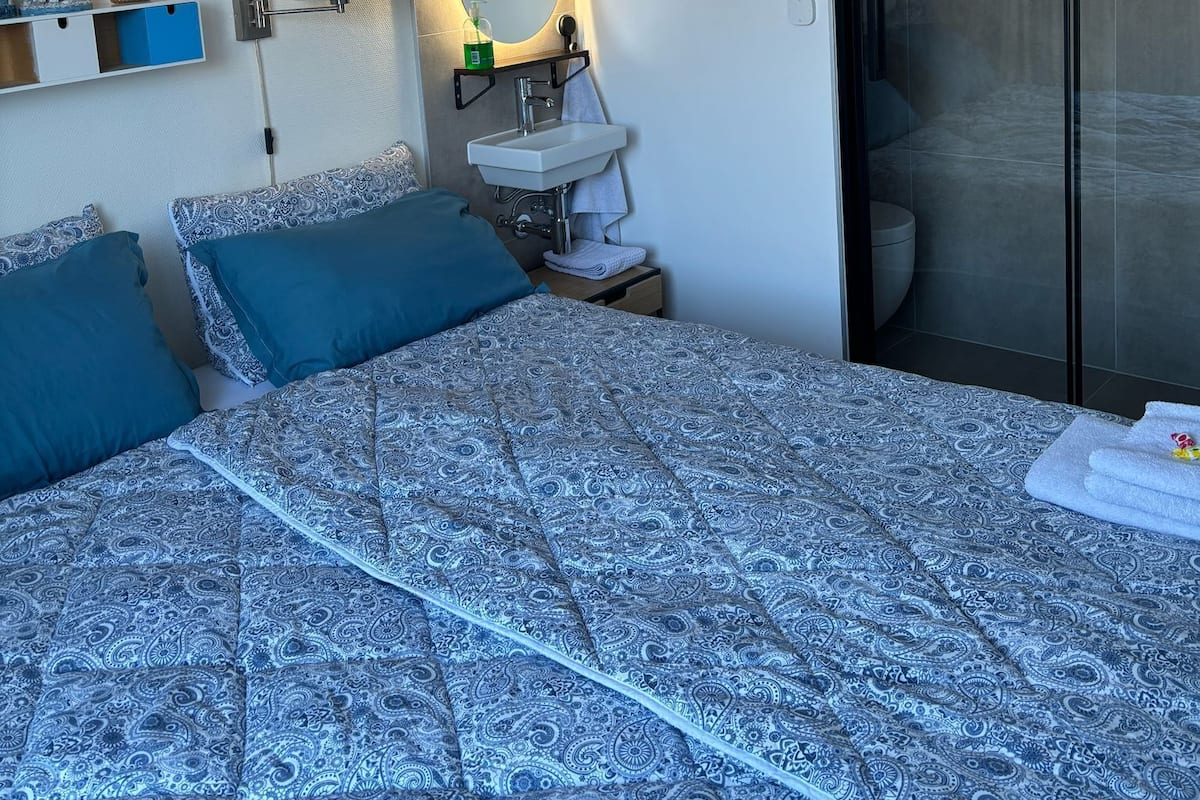 A modern bedroom with a patterned bedspread, a bedside table with a lamp, a wall-mounted mirror, and a bathroom visible through a glass door.