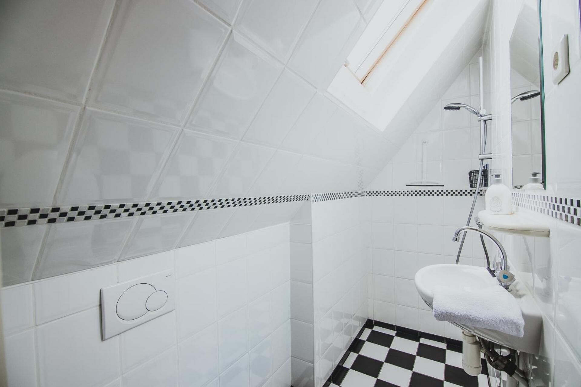 A small, modern bathroom with white tiles, a checkered floor, a towel on the sink, and a window above the shower.