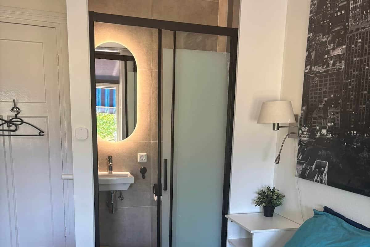 A bedroom with a bed, a nightstand, a wall-mounted lamp, and a bathroom visible through a glass door.