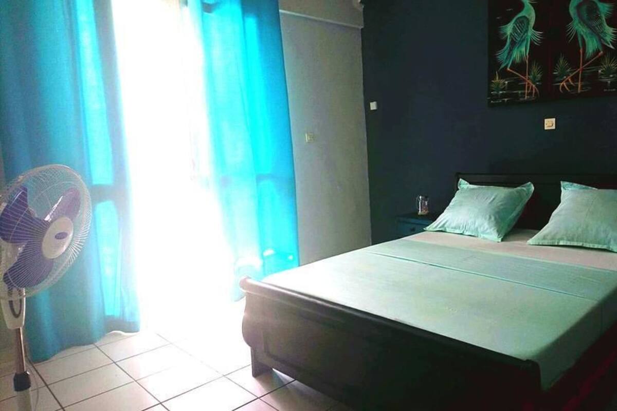 A bedroom with a bed, two pillows, a nightstand, a wall-mounted artwork, and a standing fan.