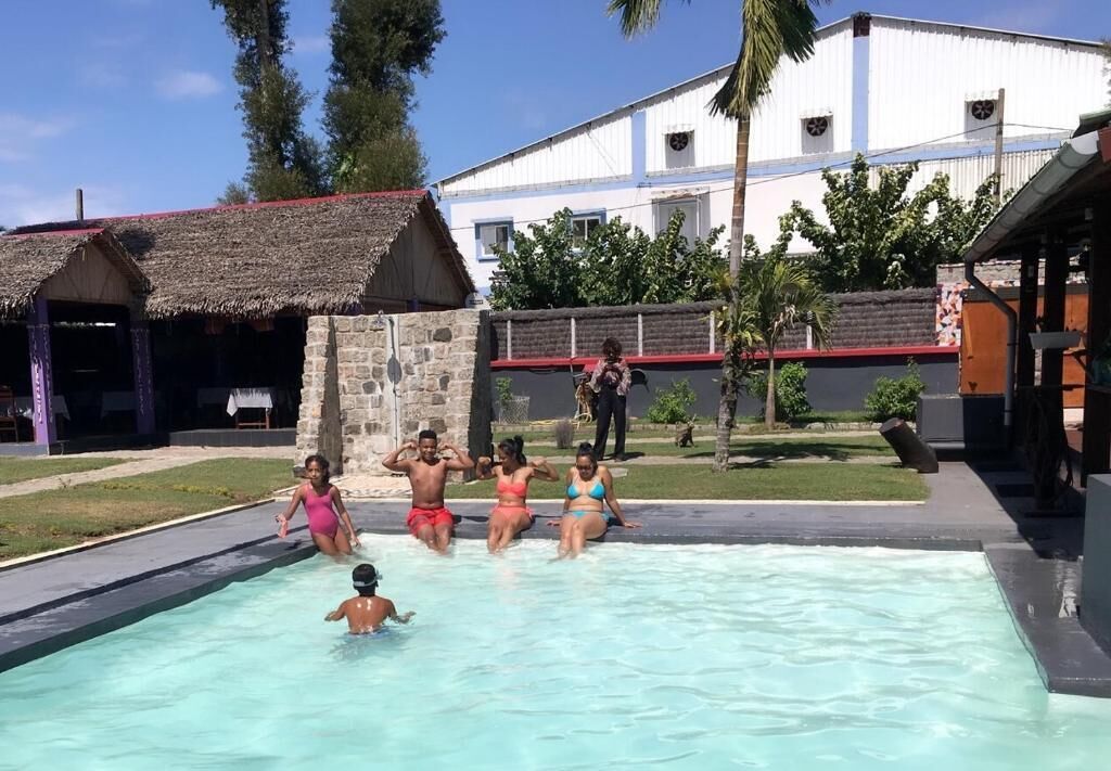 A swimming pool area with a thatched-roof structure, a stone wall, and a building with a red roof in the background.