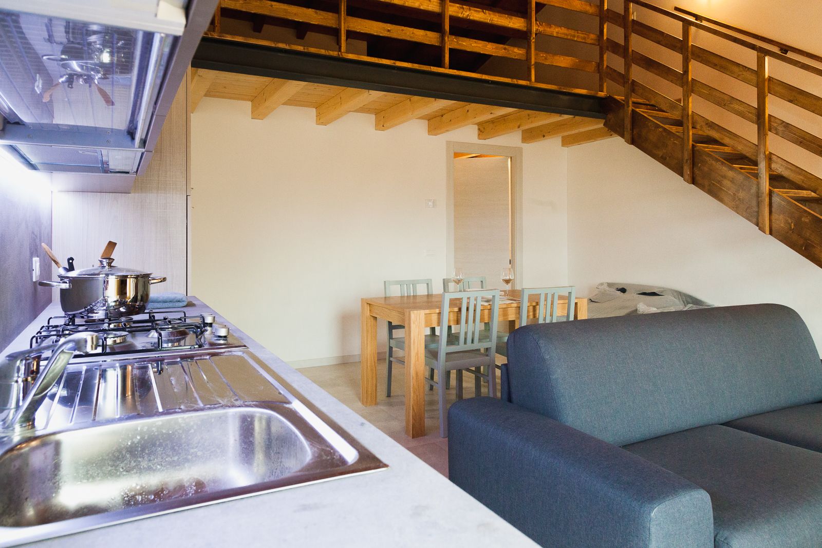 Une cuisine compacte équipée d’une cuisinière, d’un évier et de poutres en bois au plafond. On aperçoit à l’arrière-plan un coin repas avec une table et des chaises.
