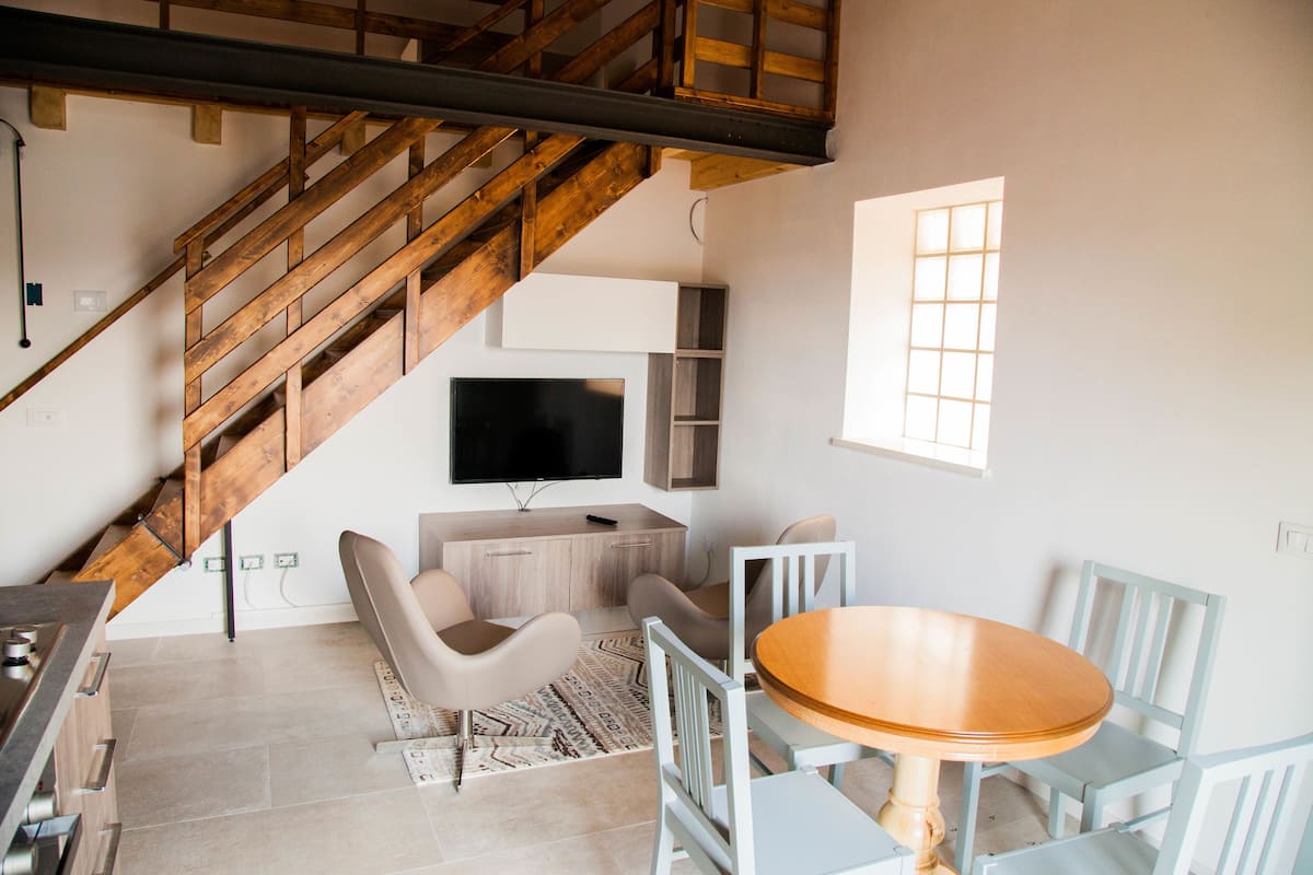 Ein modernes Wohnzimmer mit einer Holztreppe, einem Flachbildfernseher, einem Esstisch mit Stühlen und einem gemusterten Teppich.