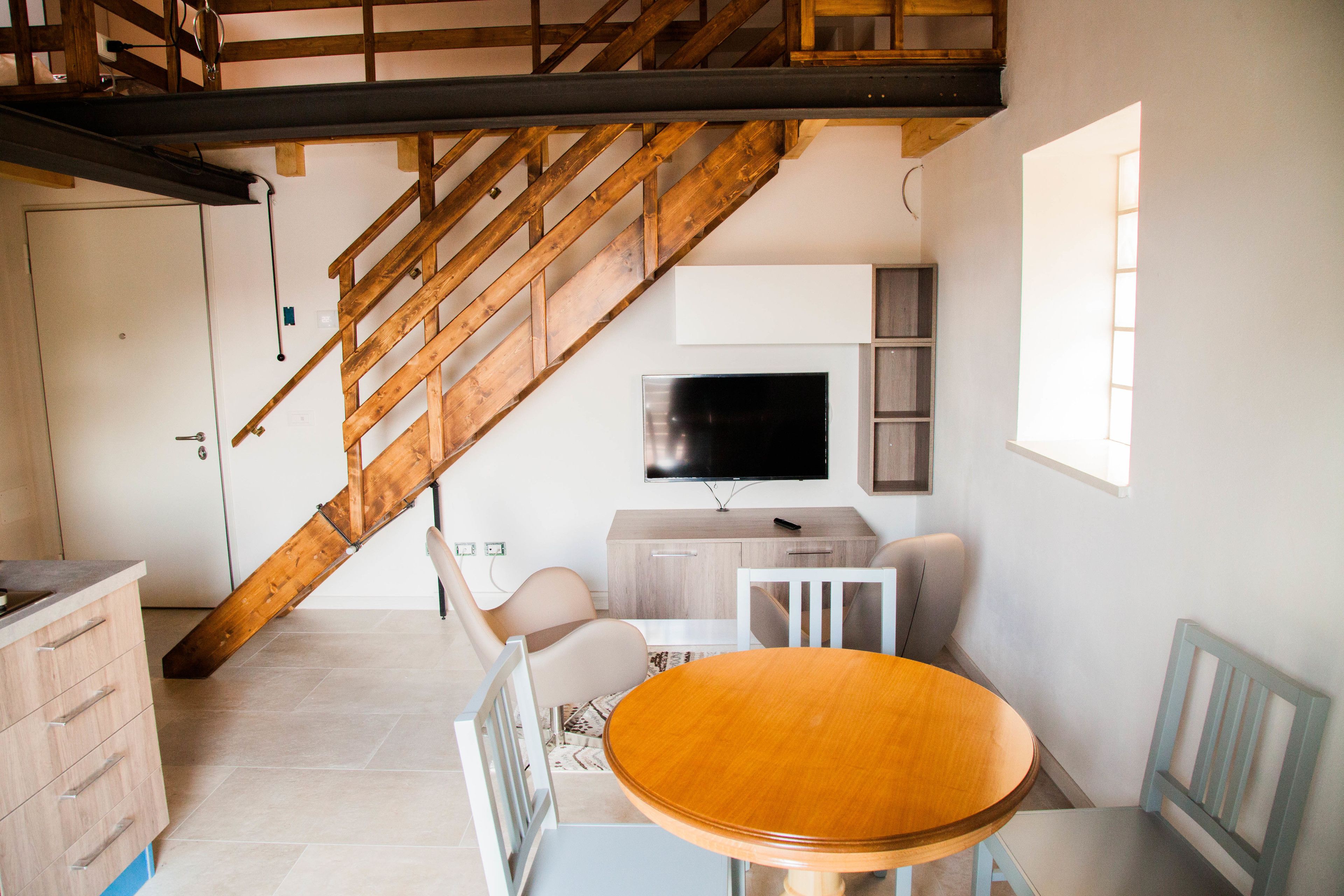 A modern living room with a wooden staircase, a flat-screen TV, a dining table, and a large window.