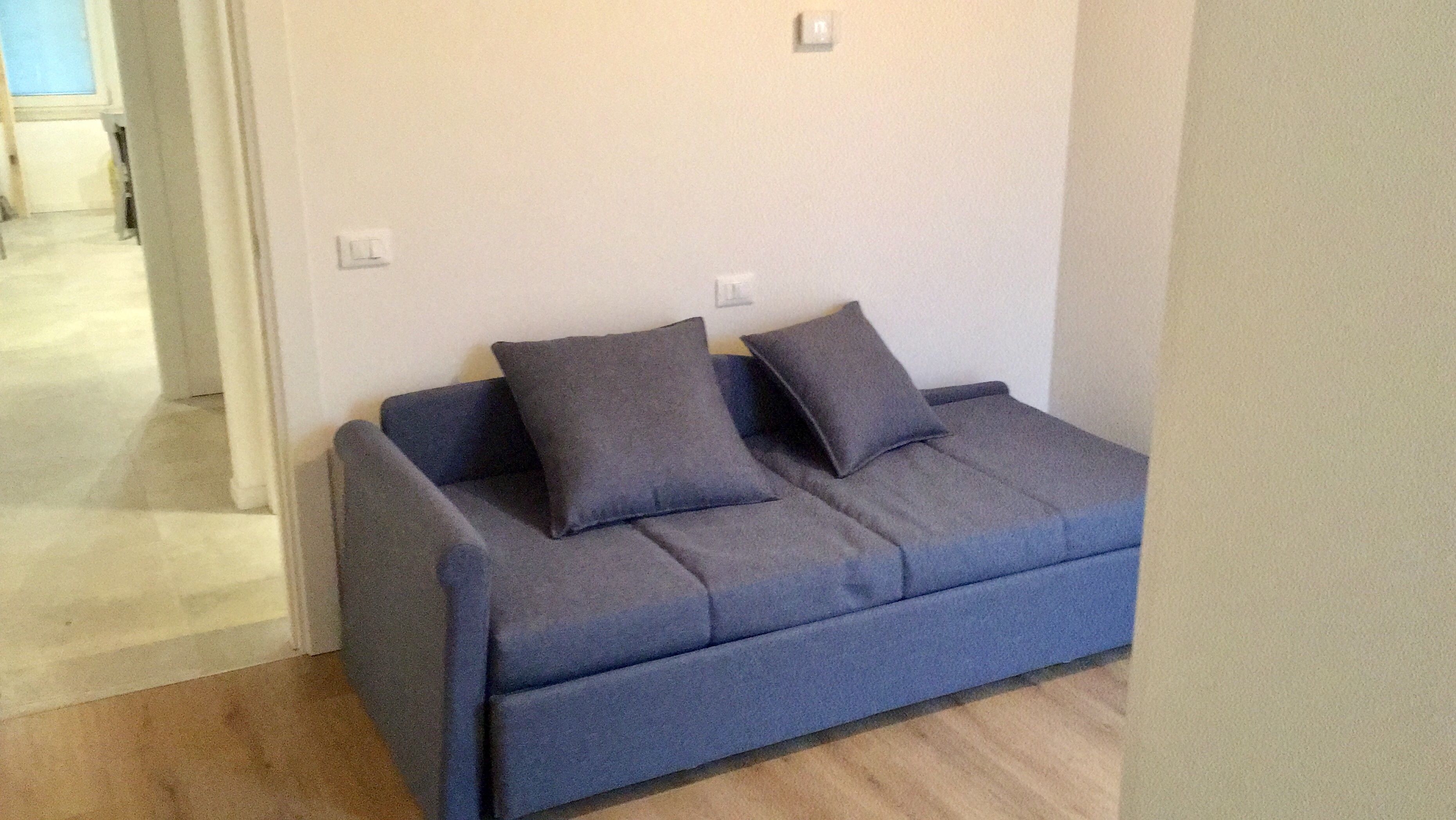 Un petit canapé de couleur sombre, agrémenté de deux coussins, est placé contre un mur uni. À proximité, on aperçoit un interrupteur et une porte qui donne sur une autre pièce.
