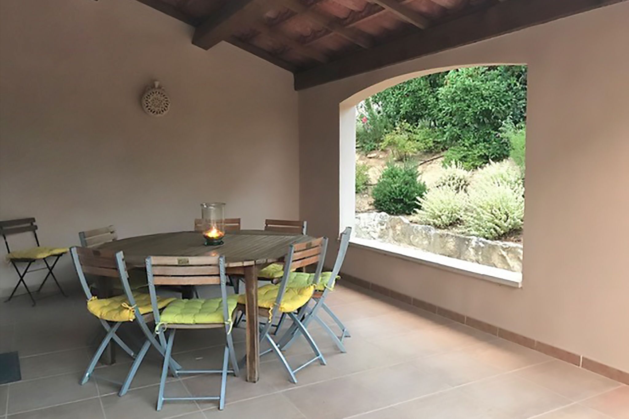 Una mesa redonda de madera con seis sillas, cada una con un cojín amarillo, en un espacio exterior cubierto que ofrece vistas a zonas verdes a través de una ventana.