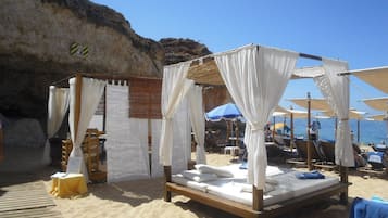 Een strandhut met een houten bed en een witte luifel, omringd door strandstoelen en parasols.