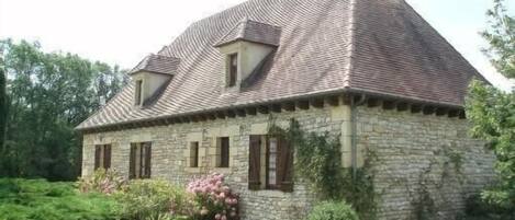 A stone house with a pitched roof, a chimney, and a small garden with pink flowers.