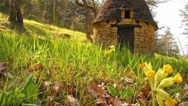 Ein Steinhäuschen mit kegelförmigem Dach, umgeben von üppigem Grün und gelben Blüten.