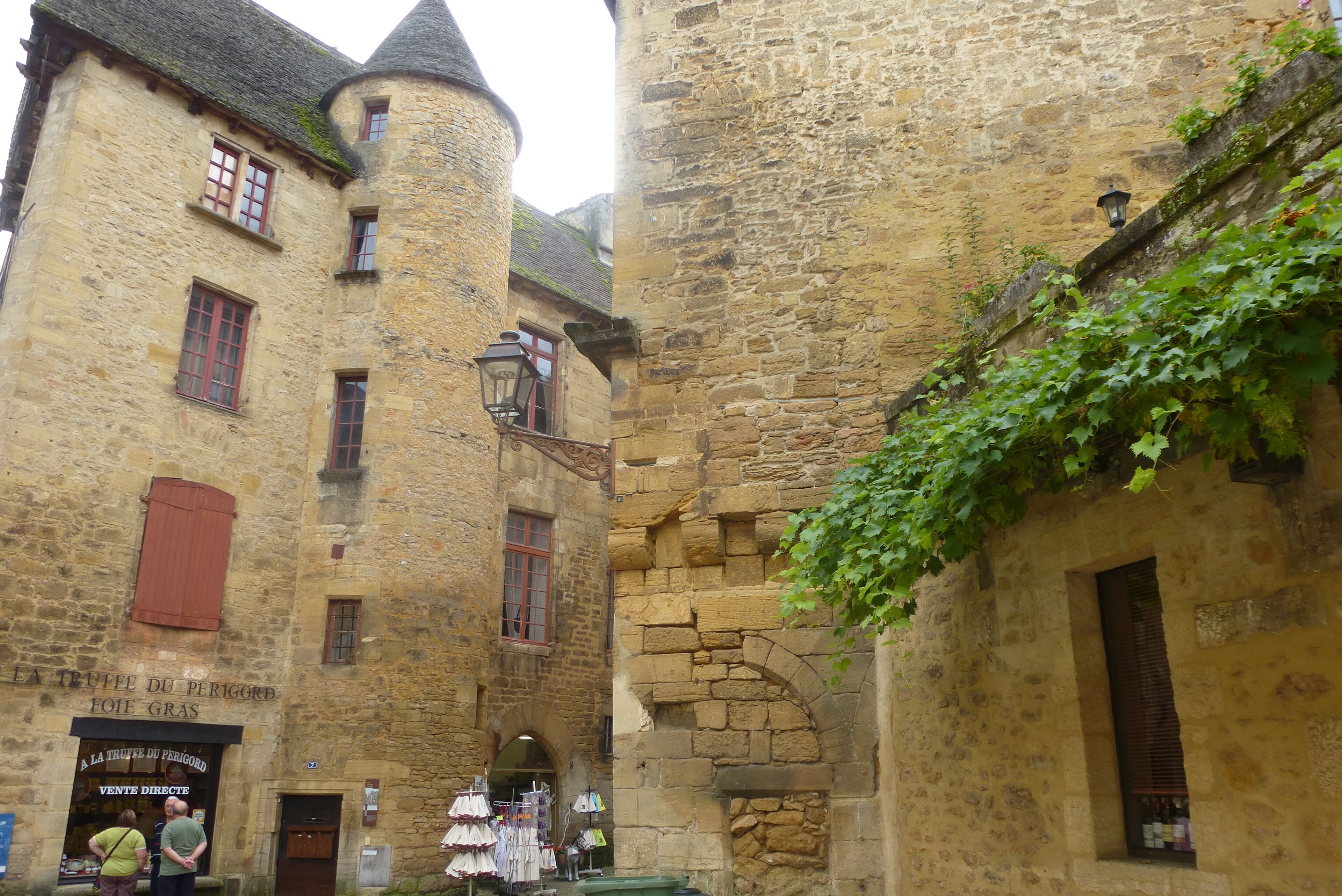 A narrow medieval street with stone buildings, a shop with clothing items displayed outside, and a sign indicating a direct sale.
