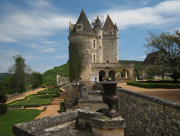 A historic castle with a central tower, a well-maintained garden, and a stone bridge leading to the entrance.