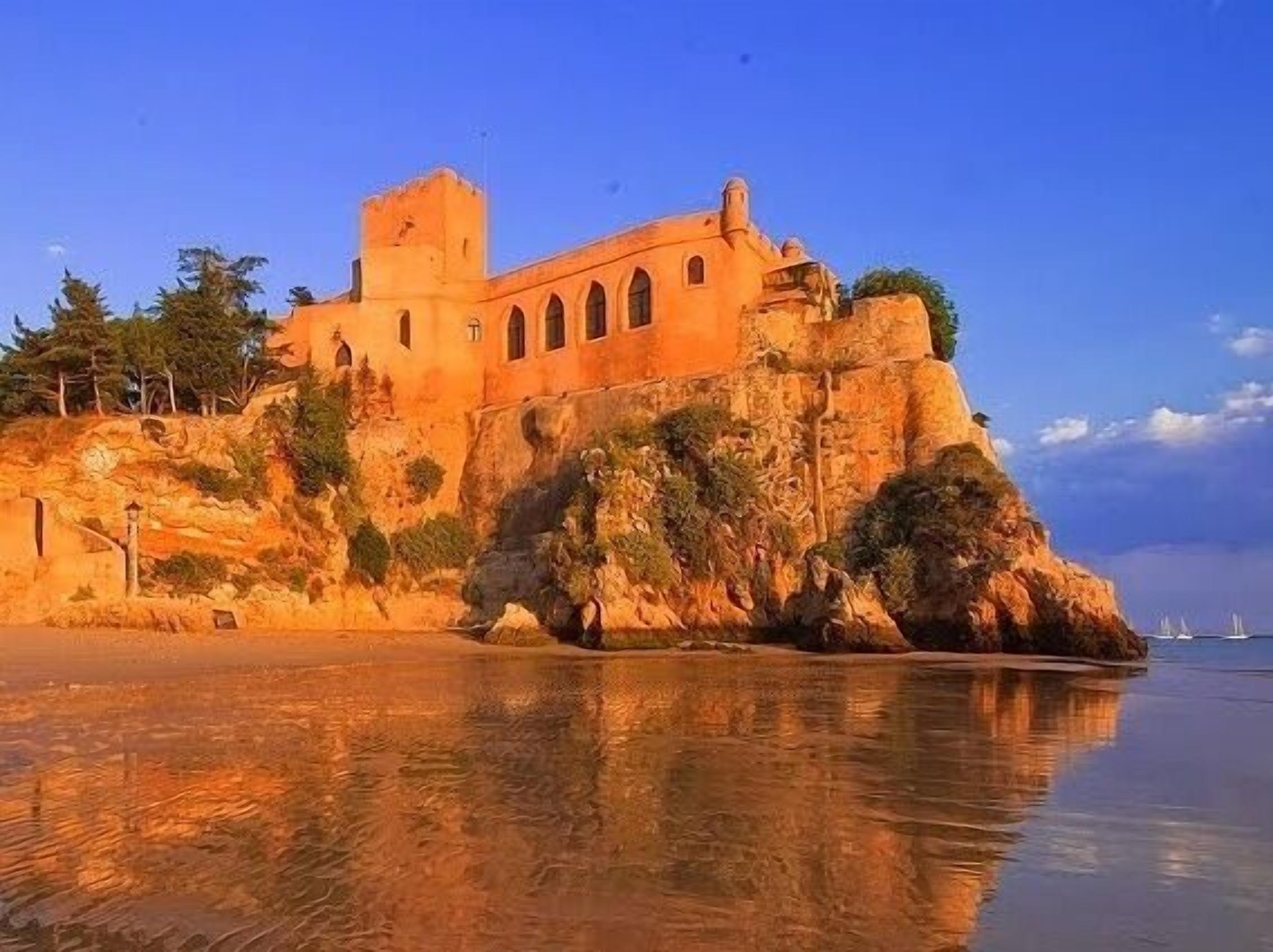 Un castell costaner amb una torre destacada, ubicat en una roca amb una vista clara del mar.