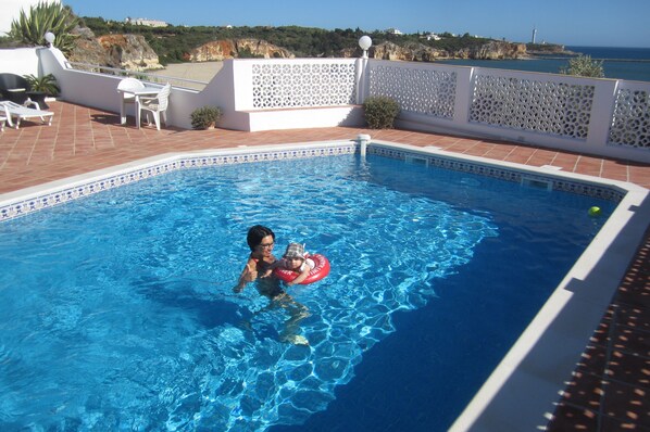 A swimming pool with clear blue water, a child in a red float, and a view of the sea and hills in the background.