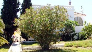 Un edificio con fachada blanca, tejado de tejas rojas y una pequeña torre. Cuenta con un jardín bien cuidado con diversas plantas y un sendero que conduce al edificio.