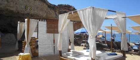 Un cabanon de plage doté d’un lit en bois et d’un auvent blanc, entouré de chaises de plage et de parasols.