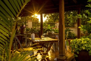 Un espace extérieur aménagé en terrasse, avec des tables et des chaises en bois, entouré de plantes en pot et d’une clôture.