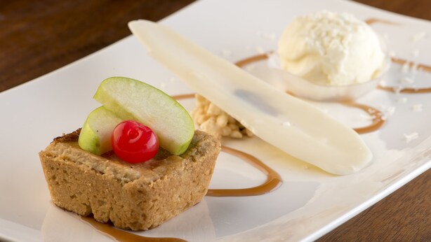 Een nagerechtenbord met een plak cake, gegarneerd met een kers, een appelpartje en een bolletje ijs.