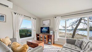 A living room with a sofa, a coffee table, and a TV on a wooden cabinet. There are large windows with curtains, a view of the outdoors, and a bookshelf. - Unique Waterfront Beach House, 1 mile from Friday Harbor (Friday Harbor)
