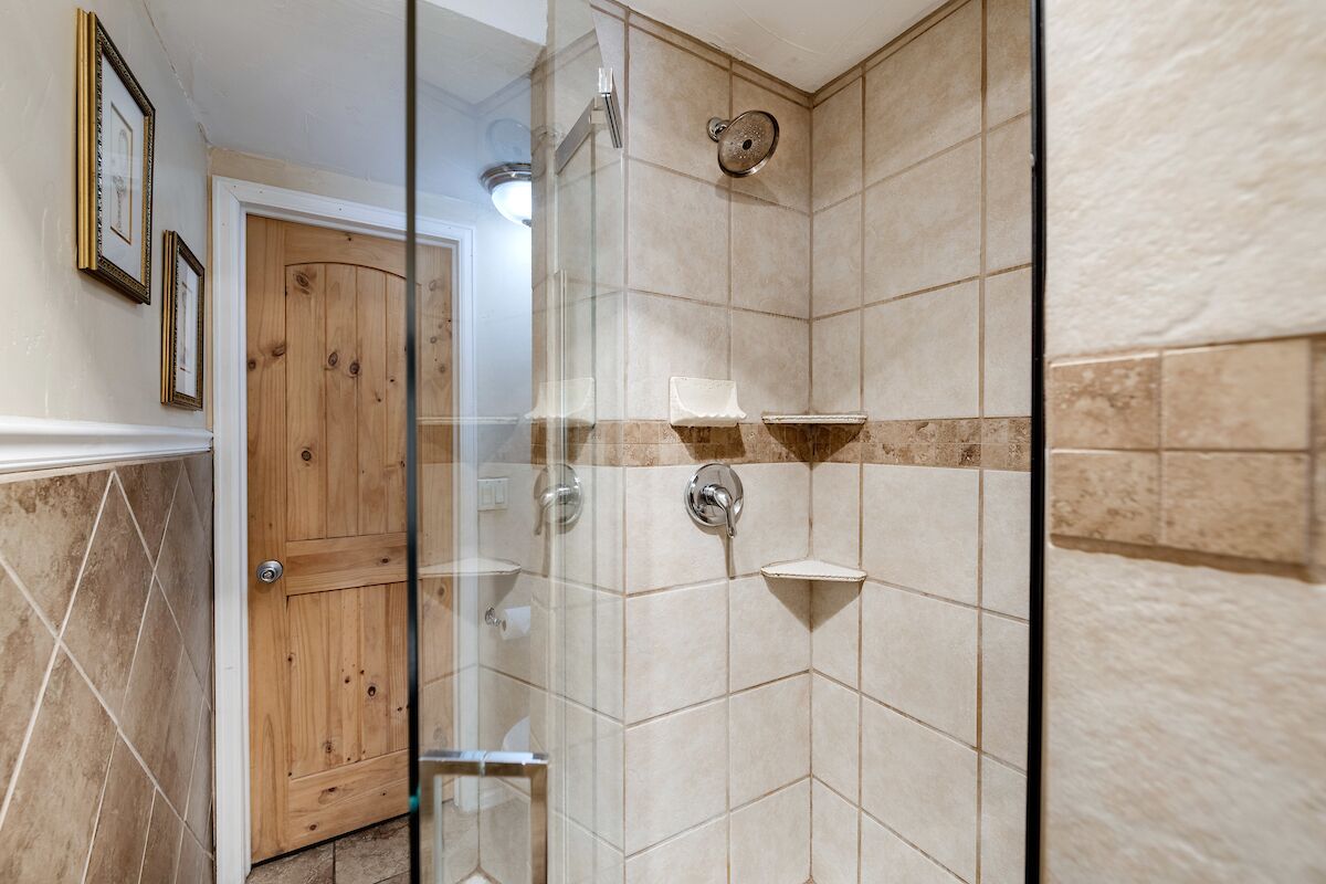 A shower with a glass door, beige tiles, and a wooden door in the background.