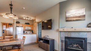 A modern kitchen with wooden cabinets, a dining area with a wooden table and chairs, a flat-screen TV mounted above the fireplace, and a stone fireplace. - Sunstone #329 Ski In Ski Out! 2 Bedroom 2 Bath (Mammoth Lakes)
