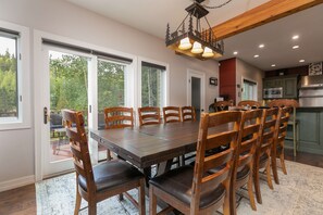 Un espace repas comprenant une grande table et des chaises en bois, un lustre et une vue sur l’extérieur grâce à de grandes fenêtres.