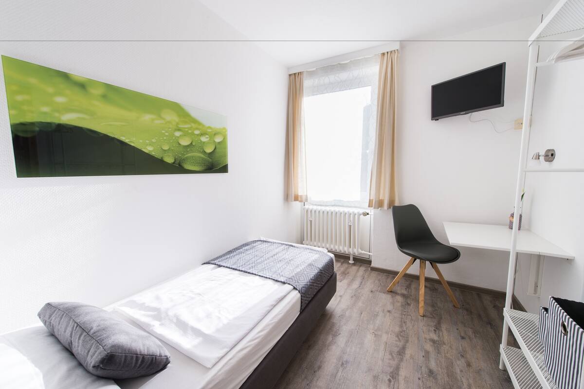 Quarto com cama, escrivaninha, cadeira, televisão e janela com cortinas.