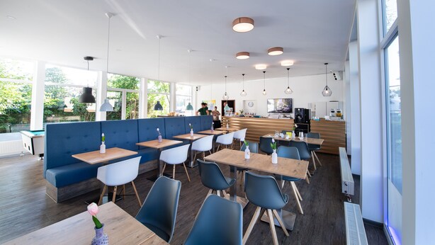 A modern restaurant with a mix of seating options including booths and tables, a kitchen area, and large windows offering views of greenery. - SOVA Hotel (Kassel)