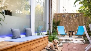A wooden bench with cushions, a dog, and blue chairs in a garden area with a wooden fence and plants. - SOVA Hotel (Kassel)
