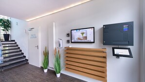 A reception area with a digital screen displaying a welcome message, a wooden reception desk, and a staircase leading to another floor. - SOVA Hotel (Kassel)