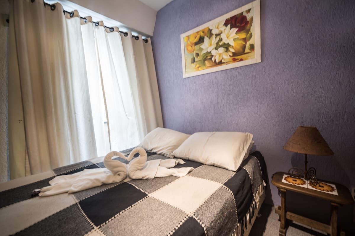 A hotel room with a checkered bedspread, a painting of flowers on the wall, a lamp on a wooden side table, and a window with curtains.
