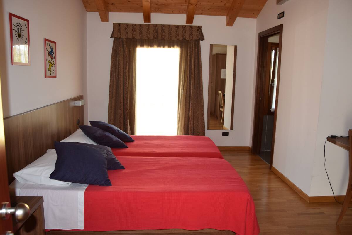 A room with two beds, wooden headboards, a large window with curtains, a mirror, and a wooden floor.