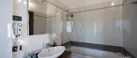 A modern bathroom with a large mirror, a white sink, a shower, and a hairdryer on the wall.