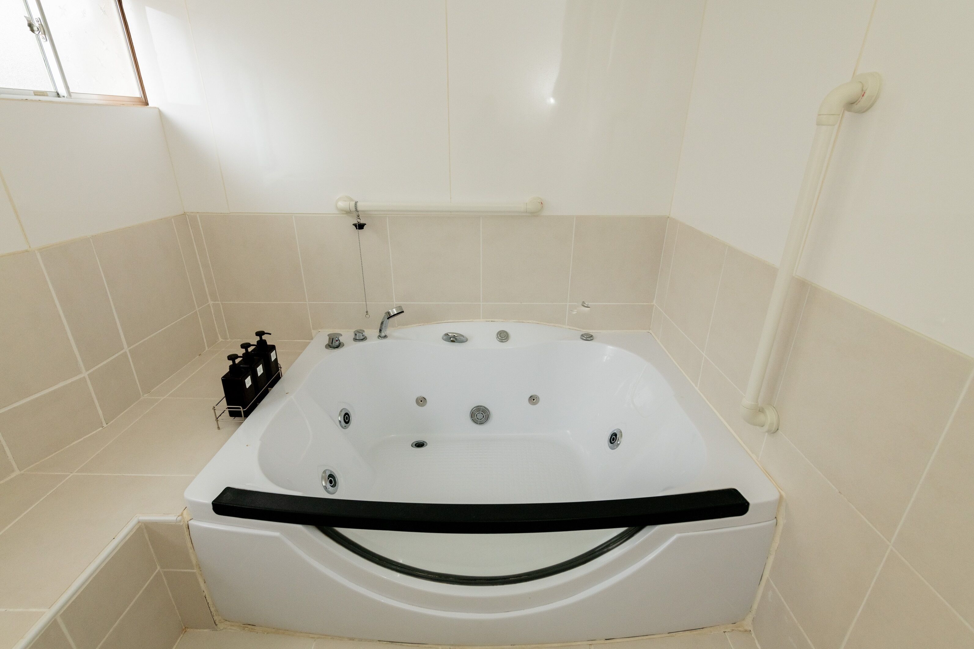 A white bathtub with a black handle, a handheld showerhead, and a black bottle of shampoo on the side.