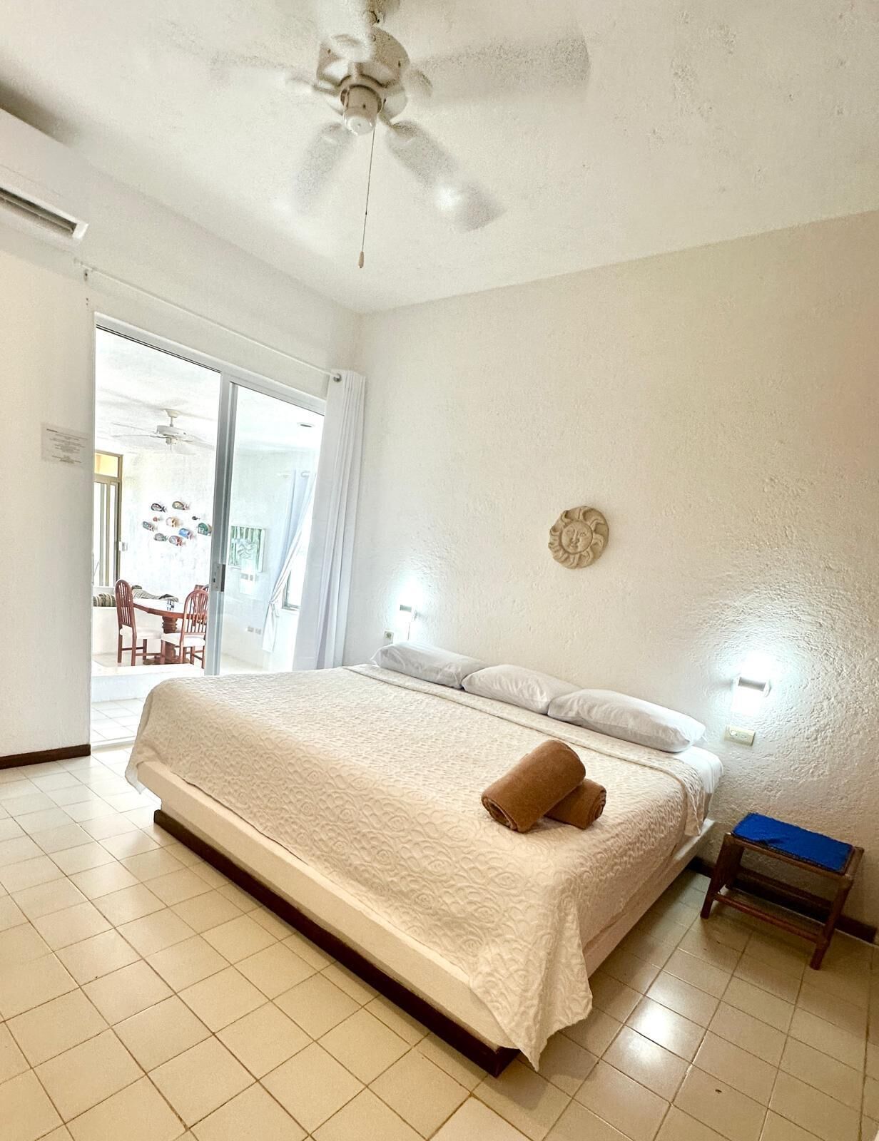Quarto com uma cama grande, um ventilador de teto, uma mesinha e uma cadeira.