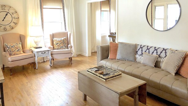 Ett väl upplyst vardagsrum med en beige soffa, ett litet bord, en stol och en klocka på väggen.