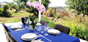 Une table à manger installée à l’extérieur, avec assiettes, couverts et verres à vin, entourée de verdure et offrant une vue sur la campagne.