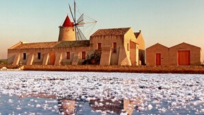 Ein Küstengebäude mit Windmühle, spiegelnder Wasseroberfläche und blauem Himmel.