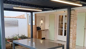 Une terrasse extérieure avec une table et des bancs en métal, un espace couvert doté d’un toit en pente, et une vue sur une maison avec une porte verte et un coin cuisine.