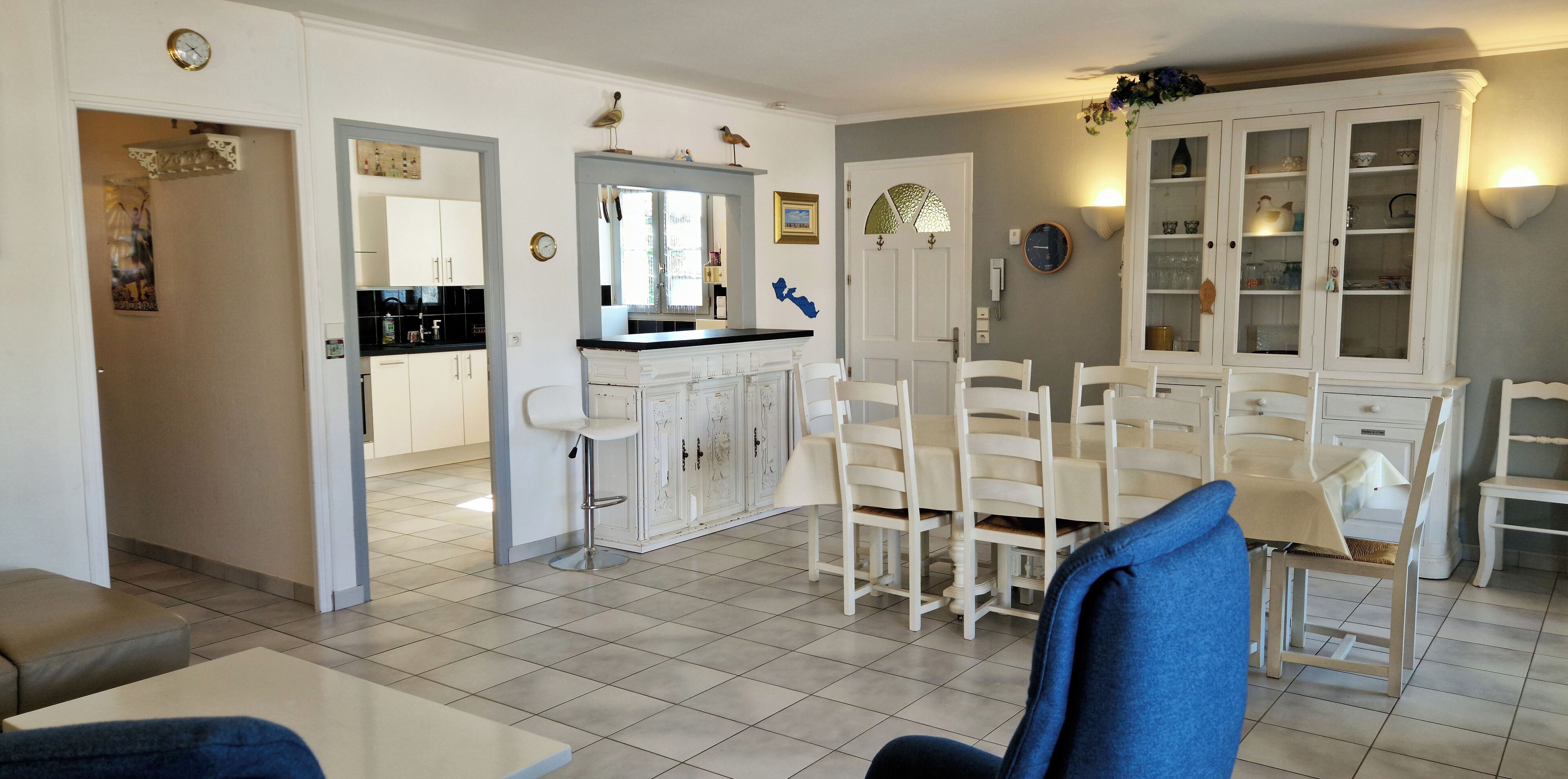 A dining area with white chairs and a table, a kitchen with white cabinets, and a clock on the wall.