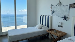 Une chambre avec une grande fenêtre, un lit recouvert d’une literie blanche, une table de chevet en bois et un filet décoratif fixé au mur, orné d’une étoile de mer.